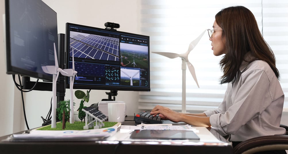 A woman with glasses is sitting at a desk working on a computer with two monitors displaying images and data related to solar panels and wind turbines. On the desk, there is a small model featuring wind turbines, solar panels, and miniature trees, along with some documents and a tablet. The background includes a window with blinds.