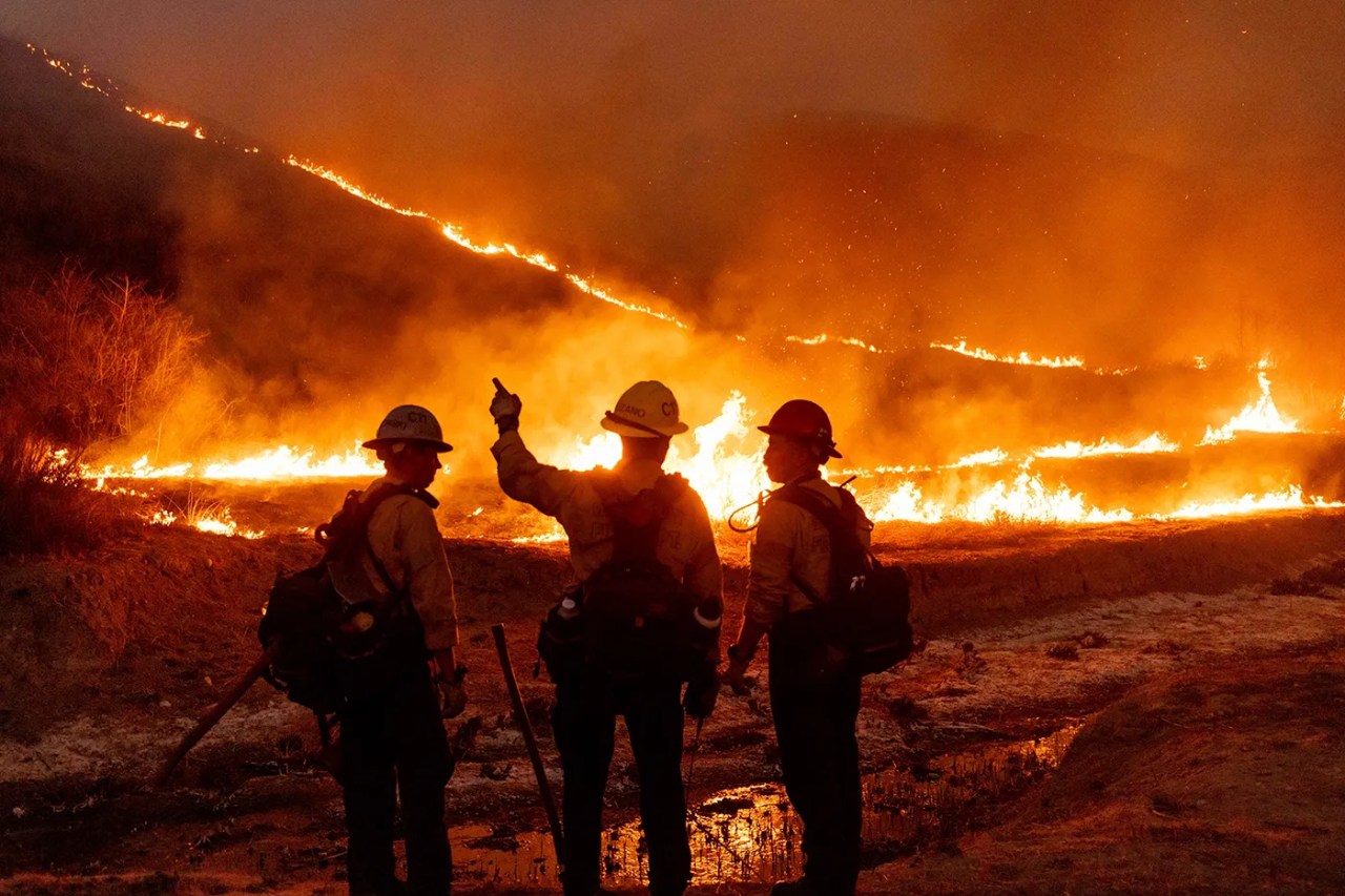 Three firefighters wearing helmets and gear stand in front of a large wildfire burning across a hillside at night, with flames and smoke illuminating the scene.