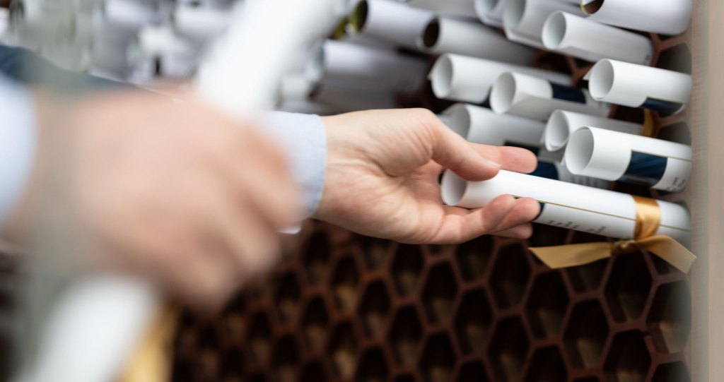 A hand is reaching to pick up a rolled-up white certificate tied with a gold ribbon from a honeycomb-style wooden rack filled with similar rolled certificates.