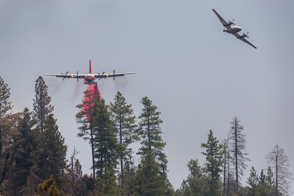 Two firefighting aircraft are flying above a forest, with one plane releasing a stream of red fire retardant over the trees below. The sky is clear with a light haze.