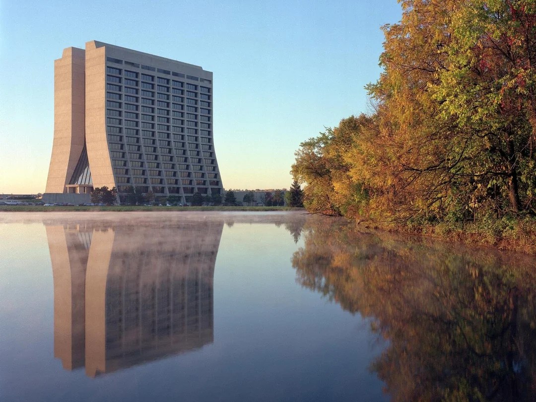 A large, modern concrete building with a unique curved design stands near a calm body of water, reflecting its image. To the right, there are trees with autumn foliage in shades of yellow, orange, and green, also reflected in the water. The sky is clear and blue, suggesting a peaceful, early morning or late afternoon scene.