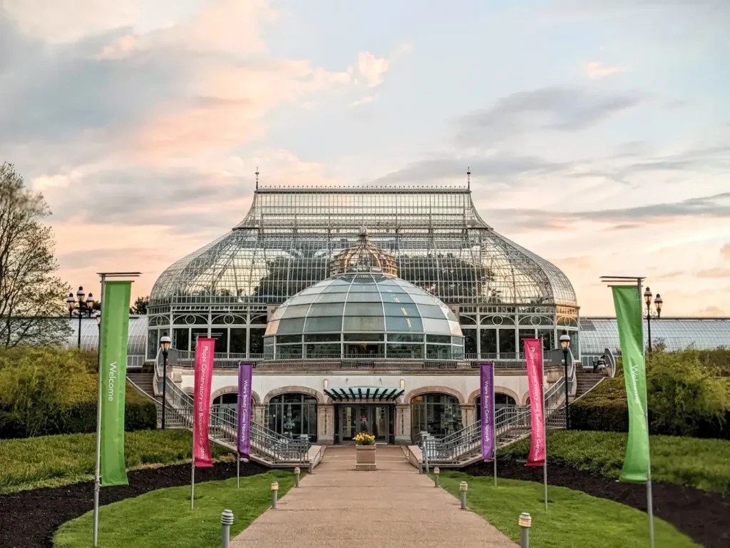 A large glass conservatory building with a domed roof and intricate metal framework, set against a partly cloudy sky at sunset. A paved walkway leads to the entrance, flanked by green and purple banners and manicured grass on either side.